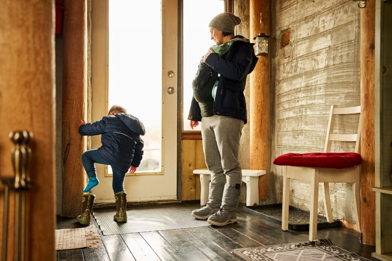 Prepping Your Plumbing for Winter. Small boy putting on his boot with his mother standing by carrying a baby boy at front door of eco cabin.